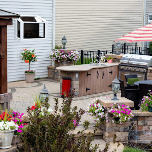 Summer flowers around an upscale brick patio with wooden gazebo, outdoor kitchen with BBQ and comfortable wicker dining furniture